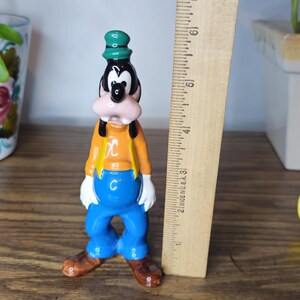 K&ouml;nnte beinhalten: Eine Vintage-Goofy-Figur, etwa 10 cm hoch, mit gr&uuml;nem Hut, orangefarbenem Hemd, blauer Hose und braunen Schuhen. Die Figur steht neben einem Holzlineal und anderen Disney-Charakterfiguren.