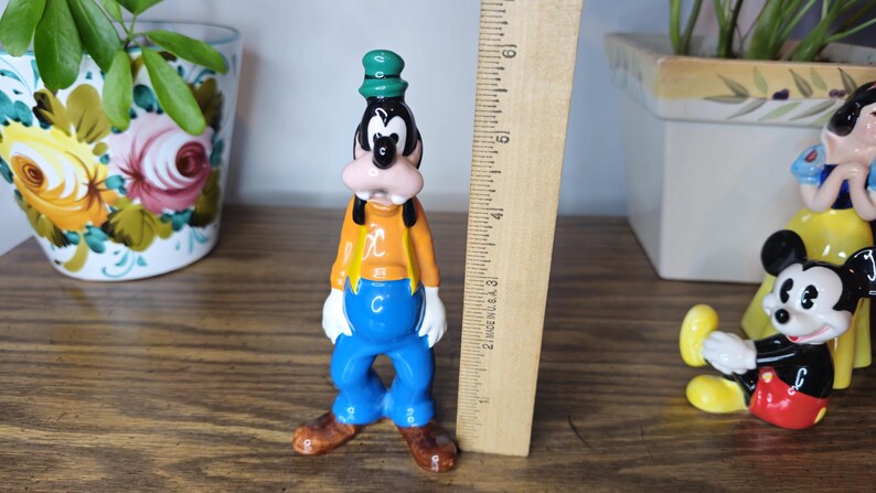 K&ouml;nnte beinhalten: Eine Vintage-Goofy-Figur, etwa 10 cm hoch, mit gr&uuml;nem Hut, orangefarbenem Hemd, blauer Hose und braunen Schuhen. Die Figur steht neben einem Holzlineal und anderen Disney-Charakterfiguren.
