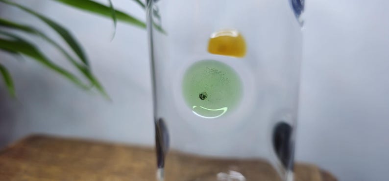 May include: A clear glass with a light green and yellow decorative elements inside. The green element has a small black dot in the center. The glass is on a wooden surface with a green plant in the background.