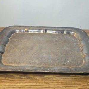 May include: An antique silver-toned rectangular tray with ornate handles. The tray has a slightly raised rim and sits on small feet. The handles feature a detailed, textured design. The tray's surface shows signs of age and use.