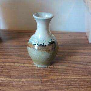 May include: A small, handcrafted ceramic vase with a white top and a green and brown body. The vase has a narrow neck and a rounded base. The colors are muted and earthy, giving it a rustic feel. It is sitting on a wooden surface.
