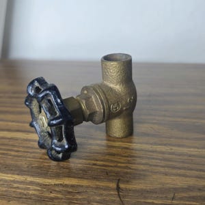 May include: A brass plumbing valve featuring a textured metallic body and a circular black handle with a scalloped edge. The valve has a vertical orientation with an opening at the top and bottom, and a horizontal stem connecting to the handle.
