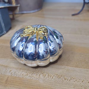 May include: A silver-plated shell-shaped jewelry box with gold-colored bow detail. The box has a blue and white floral design and measures approximately 2.5 inches in width and length.
