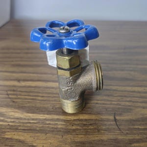 May include: A brass hose bibb valve featuring a bright blue, six-spoke circular handle at the top. The metal body has a threaded inlet at the bottom and a threaded outlet on the side, with the letters USA clearly embossed on the side of the valve.