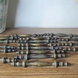 May include: A collection of seven elongated metal drawer pulls with an antique brass finish. Each pull features a central spindle design with decorative rounded accents at the ends and along the shaft, creating a uniform look for furniture hardware.
