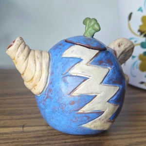 May include: A whimsical, ceramic teapot with a blue body and a white zigzag pattern. The spout and handle are beige, and a green leaf adorns the lid. The teapot is a unique decorative item.