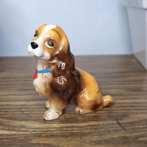 K&ouml;nnte beinhalten: Keramik-Hundefigur, braun und beige, sitzend mit blauem Halsband und rotem herzf&ouml;rmigem Anh&auml;nger. Der Hund hat ein gl&auml;nzendes Finish. Er steht auf einer Holzoberfl&auml;che.