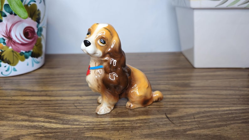 K&ouml;nnte beinhalten: Keramik-Hundefigur, braun und beige, sitzend mit blauem Halsband und rotem herzf&ouml;rmigem Anh&auml;nger. Der Hund hat ein gl&auml;nzendes Finish. Er steht auf einer Holzoberfl&auml;che.
