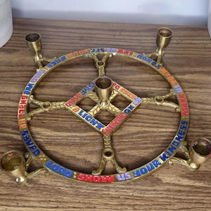 May include: A circular brass candle holder featuring four outer candle cups and one central cup. The frame is engraved with colorful text that reads: LORD SHOW US YOUR KINDNESS AND GRANT US YOUR SALVATION. A LIGHT SHINES ON US.
