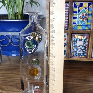 May include: A clear glass decorative bottle with colorful, round accents. The bottle is approximately 10 inches tall and has a narrow neck. The bottle is displayed on a wooden surface, with plants and decorative items in the background.