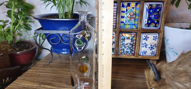 May include: A clear glass decorative bottle with colorful, round accents. The bottle is approximately 10 inches tall and has a narrow neck. The bottle is displayed on a wooden surface, with plants and decorative items in the background.