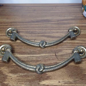 May include: Two antique brass drawer pulls with a rope design. The pulls have a curved shape with decorative circular ends and a knot in the center. They are set against a wooden surface.