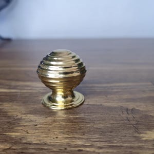 May include: A gold-colored cabinet knob with a spherical design. The knob features a series of horizontal ridges, giving it a textured appearance. The base is a simple, circular shape. The knob is likely made of metal and is designed for furniture.