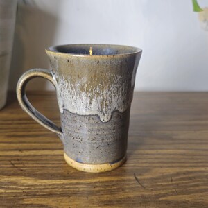 May include: A ceramic mug featuring a cylindrical body with a handle. The glaze transitions from a dark brown at the rim to a white drip pattern, ending in a speckled gray base with a tan bottom edge. The surface has a smooth, glossy finish.