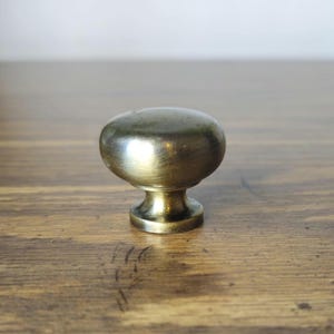 May include: A round cabinet knob with a smooth, metallic finish in a muted brass tone. The knob features a domed top surface that tapers down to a narrow, cylindrical base, providing a simple and functional design for furniture hardware.
