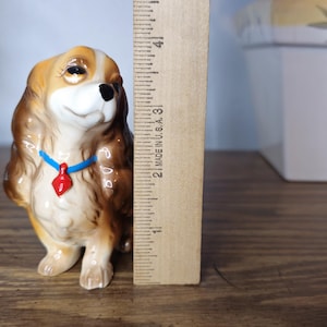 K&ouml;nnte beinhalten: Keramikfigur eines Cavalier King Charles Spaniel, ca. 7,6 cm hoch. Der Hund hat braunes und hellbraunes Fell, eine schwarze Nase und ein blaues Halsband mit rotem Anh&auml;nger. Die Figur steht auf einer Holzoberfl&auml;che.