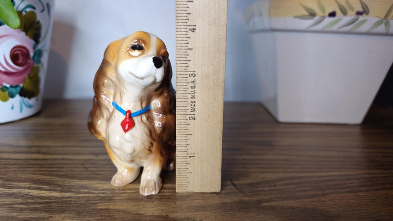 K&ouml;nnte beinhalten: Keramikfigur eines Cavalier King Charles Spaniel, ca. 7,6 cm hoch. Der Hund hat braunes und hellbraunes Fell, eine schwarze Nase und ein blaues Halsband mit rotem Anh&auml;nger. Die Figur steht auf einer Holzoberfl&auml;che.