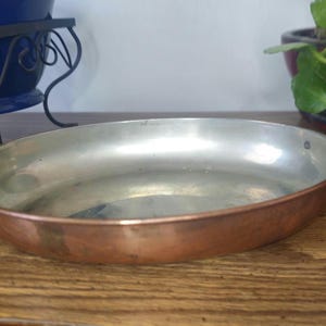 May include: Oval copper pan with brass handles. The interior is silver-colored, and the exterior is a warm copper tone. The pan sits on a wooden surface, with a blue planter and greenery in the background. A vintage kitchenware item.