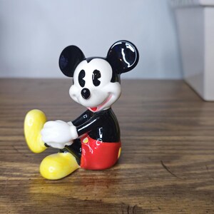 K&ouml;nnte beinhalten: Eine Keramikfigur von Mickey Mouse in sitzender Pose. Die Figur hat einen schwarzen K&ouml;rper, rote Shorts, wei&szlig;e Handschuhe und gelbe Schuhe. Auf einer Holzoberfl&auml;che platziert.
