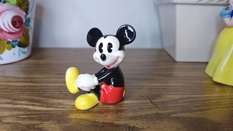 K&ouml;nnte beinhalten: Eine Keramikfigur von Mickey Mouse in sitzender Pose. Die Figur hat einen schwarzen K&ouml;rper, rote Shorts, wei&szlig;e Handschuhe und gelbe Schuhe. Auf einer Holzoberfl&auml;che platziert.