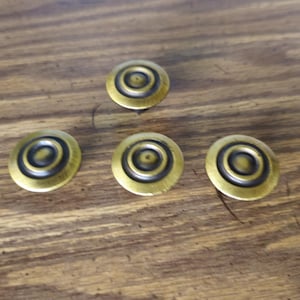 May include: Four circular metal buttons with a brushed antique brass finish. Each button features a concentric ring design with a recessed dark center and a raised outer edge, creating a layered appearance on the surface of each individual piece.