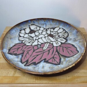May include: A ceramic plate with a floral design. The plate features a white flower with pink leaves against a blue and brown background. The plate is round and appears to be handmade.