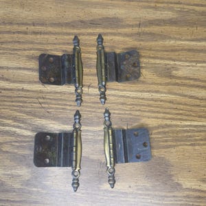 May include: Four metal cabinet hinges with a dark bronze finish and decorative finials at the top and bottom of the central pin. Each hinge features a rectangular mounting plate with two circular screw holes and a central barrel mechanism.