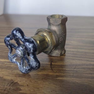 May include: A brass angle valve featuring a weathered black circular handle with a six-spoke design. The valve body is constructed from metallic brass with a threaded opening at the top and a side connection point, showing signs of age and wear.