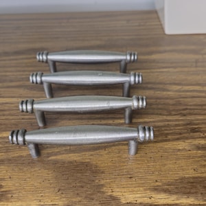 Peut inclure: Quatre poignées de placard en métal argenté disposées en rangée. Chaque poignée présente une barre centrale lisse et arrondie avec trois rainures horizontales distinctes aux deux extrémités, reliées à deux petits pieds de fixation verticaux.