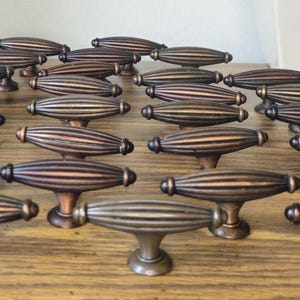 May include: A collection of numerous bronze-toned metal cabinet knobs, each featuring an elongated, oval-shaped body with vertical ribbed detailing and a central pedestal base, arranged in a grid pattern across the frame.