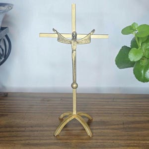 May include: A gold-colored metal crucifix with a figure of Jesus Christ. The cross has a simple design with outstretched arms. The crucifix is standing on a wooden surface. The overall theme is religious.