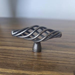 Peut inclure: Un bouton de placard en métal gris foncé présentant un design horizontal en fil torsadé en forme de cage. La quincaillerie possède un montant central relié à une base arrondie, affichant une finition texturée et industrielle sur l'ensemble.