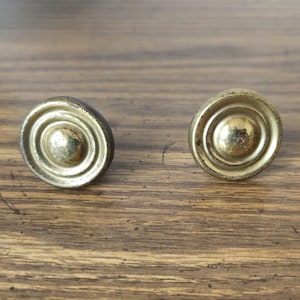 May include: A pair of antique brass cabinet knobs with a circular design. Each knob has concentric rings around a central, raised sphere. The knobs are set against a wooden surface, showcasing their aged, golden hue.