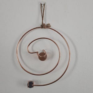 May include: Copper wire spiral ornament with a small brown bead at the center and a larger one at the end. The ornament is suspended by a natural fiber cord and is hanging against a white wall.