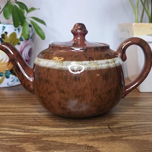 May include: Brown ceramic teapot with a white and green stripe around the middle. The teapot has a curved handle and a lid with a small knob. The teapot is sitting on a wooden surface.