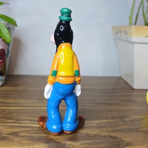 K&ouml;nnte beinhalten: Eine Keramikfigur von Goofy, gekleidet in einer gelben Jacke, blauen Hose und einem gr&uuml;nen Hut. Die Figur ist Teil einer Disney-Sammlung, mit anderen Figuren von Mickey Mouse und Schneewittchen im Hintergrund.