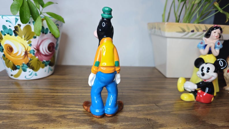 K&ouml;nnte beinhalten: Eine Keramikfigur von Goofy, gekleidet in einer gelben Jacke, blauen Hose und einem gr&uuml;nen Hut. Die Figur ist Teil einer Disney-Sammlung, mit anderen Figuren von Mickey Mouse und Schneewittchen im Hintergrund.