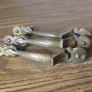 May include: Three brass drawer pulls with a curved, elongated handle design and decorative circular mounting plates at each end. The metal has a tarnished, aged finish with visible wear and patina, featuring a central grip section.