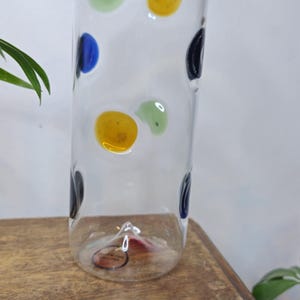 May include: A clear glass vase with colorful polka dots in blue, yellow, green, and black. The vase is cylindrical and sits on a wooden surface, with green plants in the background.