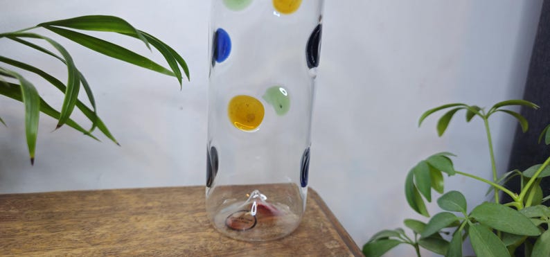 May include: A clear glass vase with colorful polka dots in blue, yellow, green, and black. The vase is cylindrical and sits on a wooden surface, with green plants in the background.