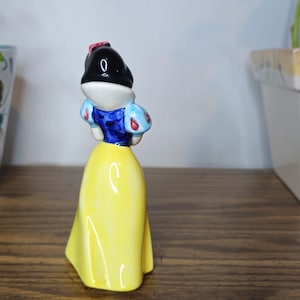 K&ouml;nnte beinhalten: Eine Keramikfigur von Schneewittchen, mit schwarzem Bob, wei&szlig;er Haut und gelbem Kleid. Die Figur hat ein blaues Oberteil mit roten und wei&szlig;en Akzenten. Die Figur steht auf einer Holzoberfl&auml;che.