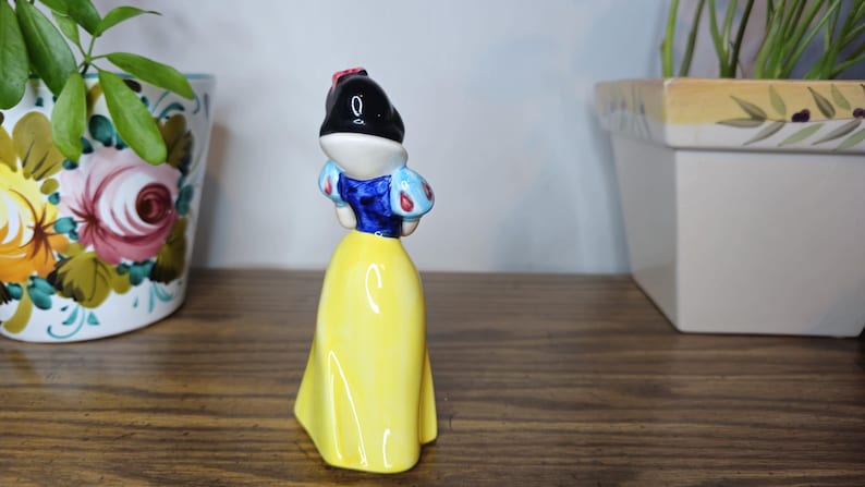 K&ouml;nnte beinhalten: Eine Keramikfigur von Schneewittchen, mit schwarzem Bob, wei&szlig;er Haut und gelbem Kleid. Die Figur hat ein blaues Oberteil mit roten und wei&szlig;en Akzenten. Die Figur steht auf einer Holzoberfl&auml;che.