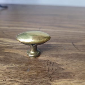 May include: A brass cabinet knob with a mushroom-shaped top and a short, cylindrical base. The knob has a slightly aged appearance, with visible wear and a warm, golden hue. It sits on a wooden surface.