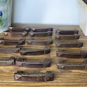 May include: A collection of twelve bronze-toned metal drawer pulls arranged in rows. Each handle features a curved bar design with a small, raised pyramid-shaped detail at both ends, providing a consistent look for cabinetry or furniture hardware.