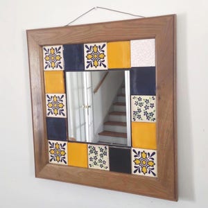 May include: Square mirror with a wooden frame. The frame is decorated with colorful tiles in yellow, navy blue, white, and floral patterns. The mirror hangs on a white wall.