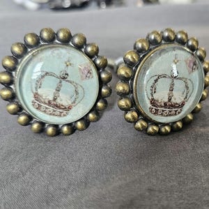 May include: Two antique-style cabinet knobs with a crown design. The knobs have a bronze-colored beaded edge and a light blue background. The crown is detailed with brown and white accents.