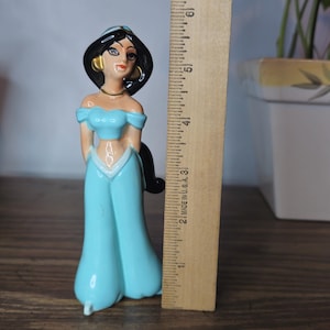 K&ouml;nnte beinhalten: Eine Keramikfigur von Prinzessin Jasmin aus Aladdin, die ein t&uuml;rkisfarbenes schulterfreies Oberteil und eine passende Hose tr&auml;gt. Die Figur ist etwa 13 cm gro&szlig;. Ein Holzlineal befindet sich neben der Figur.