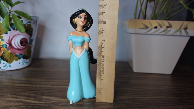 K&ouml;nnte beinhalten: Eine Keramikfigur von Prinzessin Jasmin aus Aladdin, die ein t&uuml;rkisfarbenes schulterfreies Oberteil und eine passende Hose tr&auml;gt. Die Figur ist etwa 13 cm gro&szlig;. Ein Holzlineal befindet sich neben der Figur.