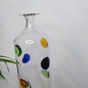 May include: A clear glass vase with a slender neck and a wide base. The vase is decorated with colorful, irregularly shaped polka dots in yellow, green, blue, and black. The vase is displayed against a white background with green plants.