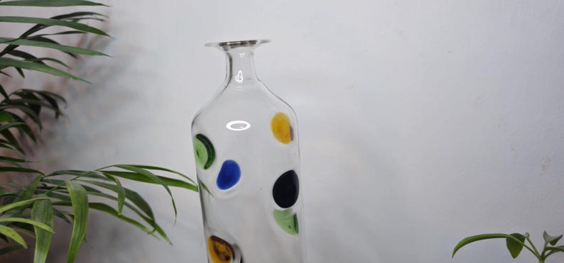 May include: A clear glass vase with a slender neck and a wide base. The vase is decorated with colorful, irregularly shaped polka dots in yellow, green, blue, and black. The vase is displayed against a white background with green plants.
